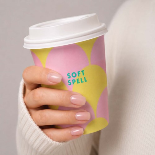 En hånd holder en pink og gul papirkop med hvid låg, mærket Soft Spell.