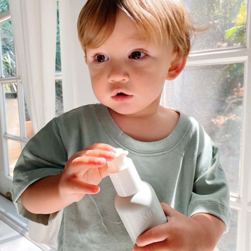 Een jonge jongen in een groen shirt houdt een witte fles en kijkt nieuwsgierig, naast een raam met zonlicht.