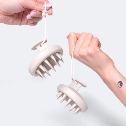 Twee handen houden witte massagetools met punten, hangend aan touwen, tegen een eenvoudige achtergrond.