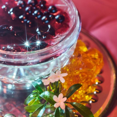 Een glas met bessen drink met zwarte bessen bovenop, gele gelei en groene bladeren met roze bloemen.