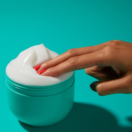 Eine Hand taucht die Finger in ein weißes Cremeglas vor einem blauen Hintergrund.