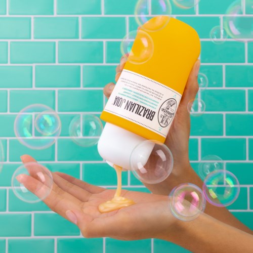 En person häller lotion från en gul flaska på sin hand, med bubblor som flyter runt mot en turkos kaklad bakgrund.
