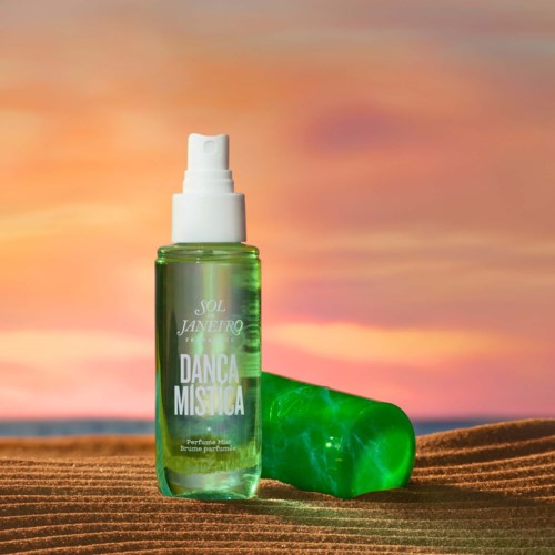 A green perfume spray bottle with a white cap, labeled 'Danca Mistica,' on a sandy beach at sunset.