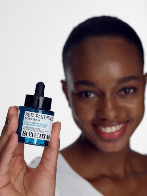 Eine lächelnde Frau hält eine blaue Tropfflasche mit den Labels 'SOME BY' und 'BETA PANTHENOL REPAIR SERUM'.