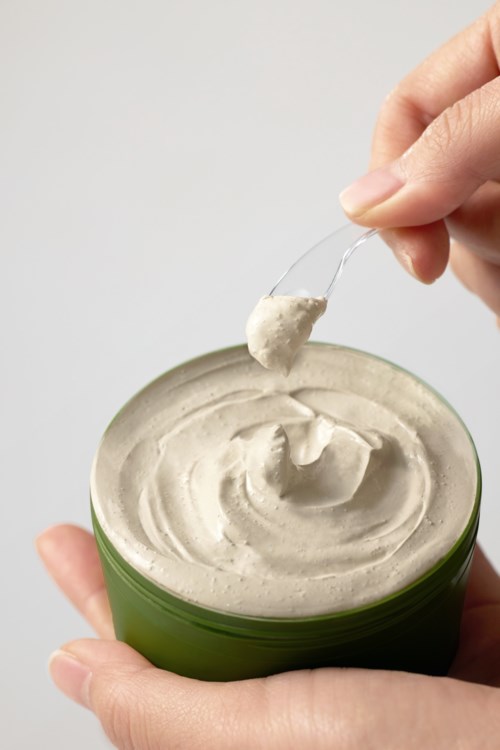 En person doppar en sked i en grön behållare fylld med krämigt, beige ämne, troligen en mat- eller kosmetikprodukt.