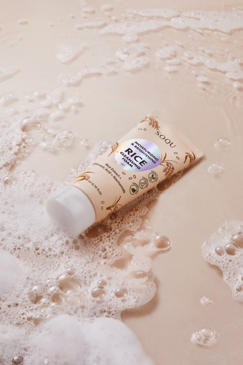 En tub med risbaserad hudvårdsprodukt flyter i skum på en beige yta.