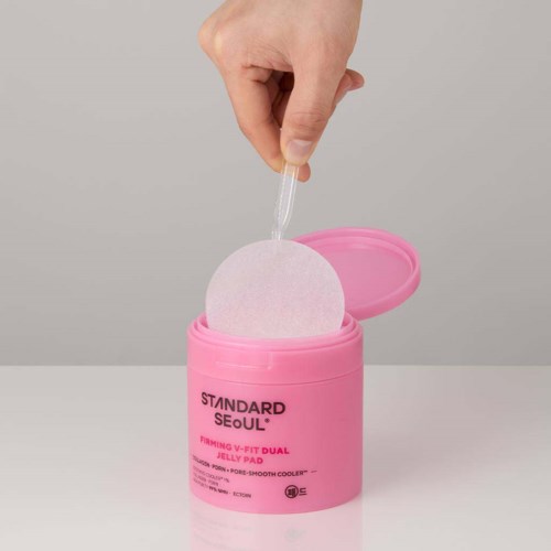 Eine Hand hebt ein weißes Reinigungsdesinfektionstuch aus einem pinken Behälter mit der Aufschrift 'STANDARD SEOUL.'