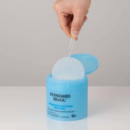 Eine Hand taucht eine Pipette in einen blauen Behälter mit der Aufschrift 'Standard Seoul', in dem sich ein Wattepad befindet.