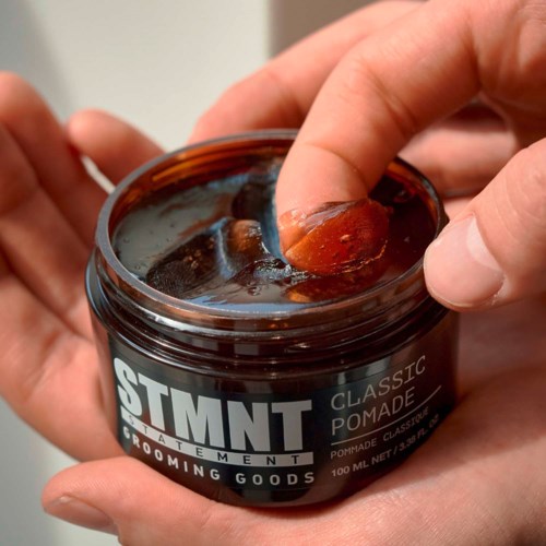 Hænder dypper fingre i en bøtte med grooming pomade mærket 'STMT Classic Pomade.' Pomaden er mørk og tyk.