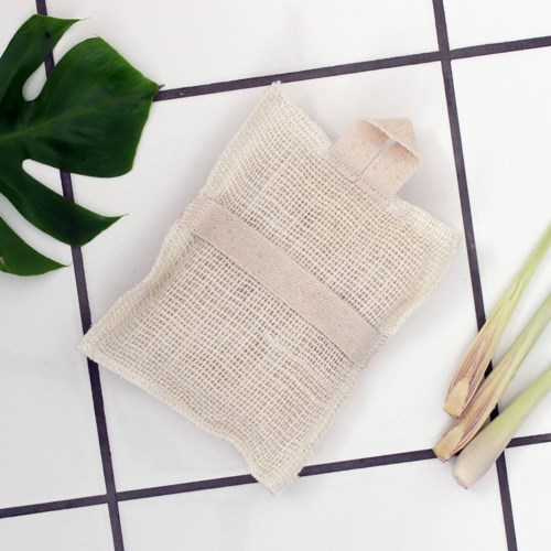 En beige tygpåse ligger på ett vitt kaklat golv med svarta foglinjer, omgivet av gröna blad och ljusfärgade växter.