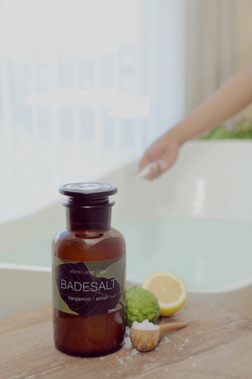 En brun flaske mærket Badesalt med citron, broccoli og en lille børste ved siden af, på en træoverflade nær et badekar.