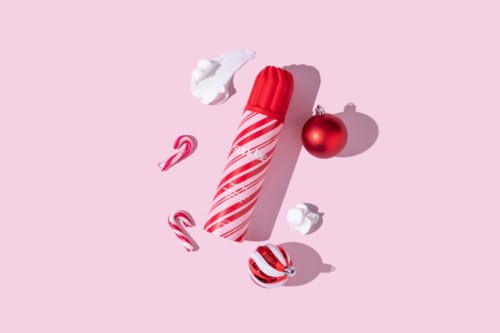 Een feestelijk kersttafereel met rode en witte peperkoekstokjes, een rode kerstbal en slagroom op roze achtergrond.