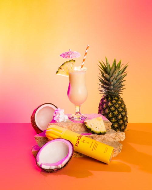 Colorful tropical scene with a pineapple, coconut, drink, and skincare products on a pink and orange background.
