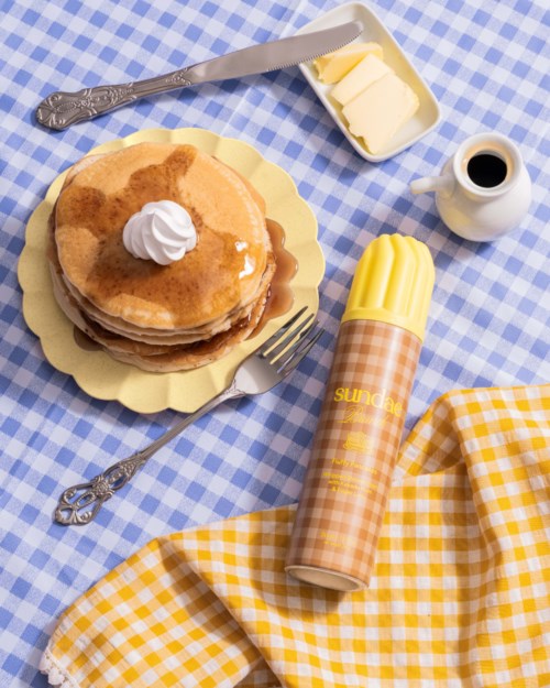 Stack av pannkakor med sirap, smör och vispgrädde på ett blått rutigt tyg.