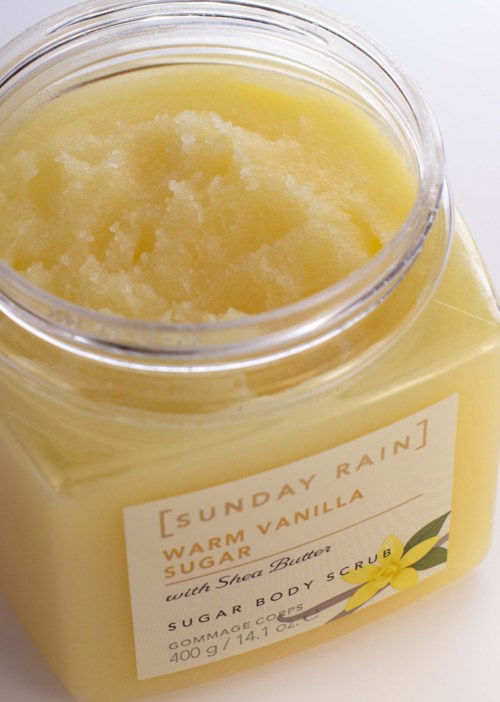 En krukke med gul kroppsskrubb med etiketten Sunday Rain Warm Vanilla Scent Scented Sugar, Sheer Butter, og 400g.