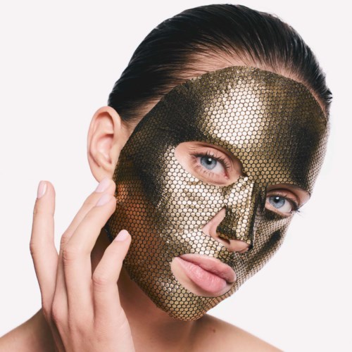 En kvinna med klarblå ögon bär en guldfärgad mask med hexagonmönster som täcker ansiktet, och rör vid sin kind med handen.