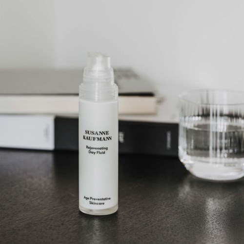 Valkoinen pullo, jossa lukee 'Rejuvenating Day Fluid' ja 'Age Preventive Skincare' mustalla pinnalla kirjojen ja vesilasillisen vieressä.