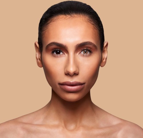 Eine Frau mit klarer Haut, braune Haare und neutralem Make-up, schaut direkt in die Kamera vor einem beigen Hintergrund.
