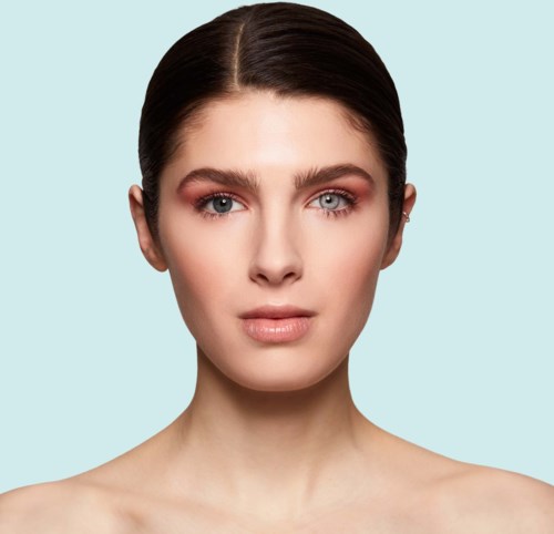Een vrouw met een lichte huid, bruin haar en natuurlijke make-up tegen een lichtblauwe achtergrond.