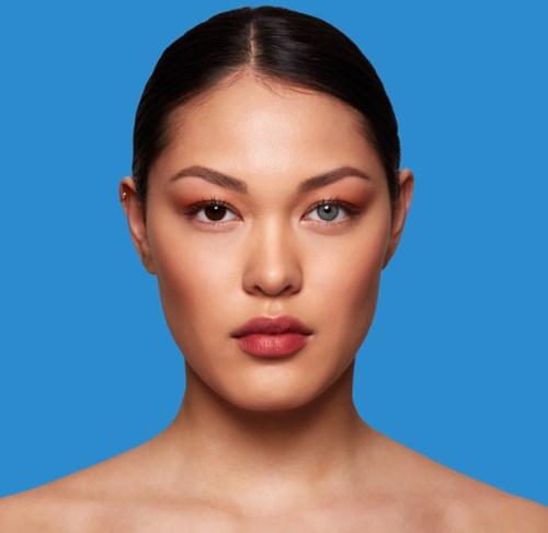 Een vrouw met een gladde huid, donker haar en één oog gedeeltelijk gesloten, tegen een blauwe achtergrond.