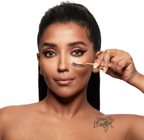 En kvinna med mörkt hår applicerar mascara på sitt öga, visar sina makeupfärdigheter och en tatuering som säger 'Stay Beautiful.'