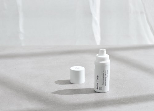 Een witte huidverzorgingsfles met dop, op een grijze ondergrond met schaduw, tegen een lichte achtergrond.