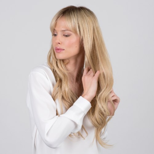 Kobieta z długimi blond włosami dotyka włosów, nosi biały top, patrzy w dół, neutralny wyraz twarzy.