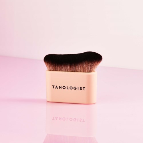 Ein Make-up-Pinsel mit schwarzen und braunen Borsten und einem cremefarbenen Griff mit "TANOLOGIST" auf rosa Hintergrund.