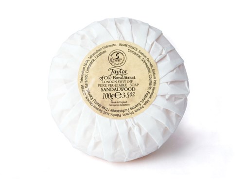 En rund tvål inlindad i vitt papper med en beige etikett som visar ingredienser, vikt och märkesdetaljer.