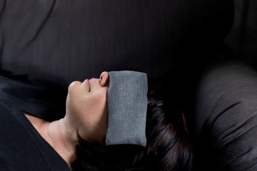 Eine Frau schläft auf einer dunklen Couch mit einem grauen Tuch über den Augen, entspannt und ruhend.