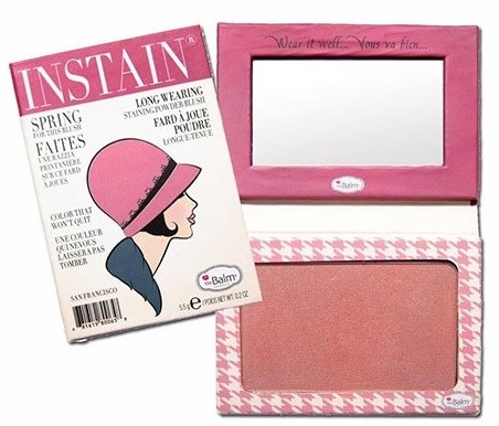 Ett rosa rouge-compact med spegel och en fyrkantig rouge inuti, samt en tidning med en kvinnahattillustration.