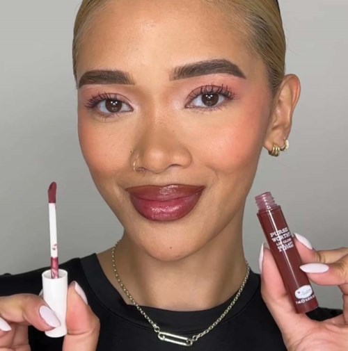 En kvinde med blondt hår, makeup, holder en læbepenegel og en læbestift, smiler til kameraet.