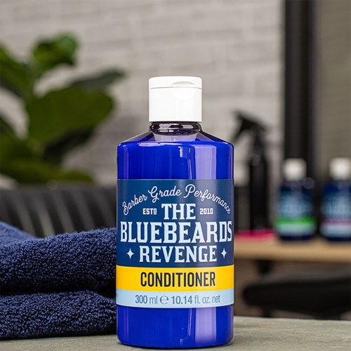 A blue bottle of conditioner labeled "The Bluebeards Revenge." Next to it is a dark towel, with a barbershop background.