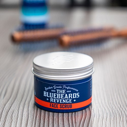 A blue and orange jar labeled "Bluebeards Revenge Face Scrub" on a wooden surface.