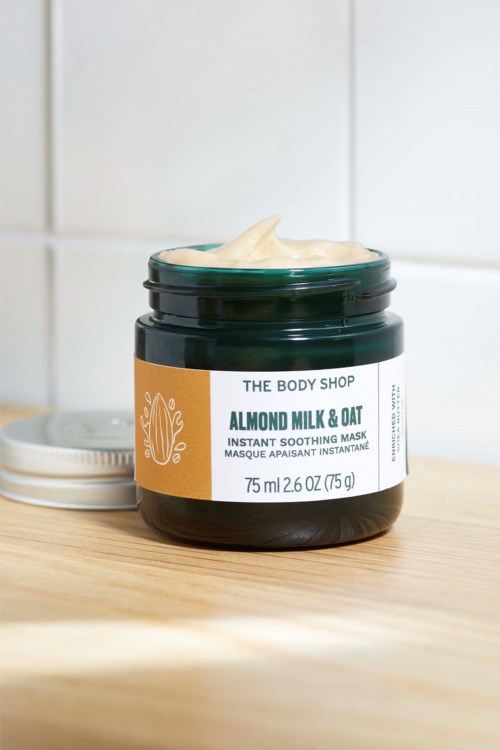 A dark jar of almond milk and oat face mask with a small amount on top, on a wooden surface in front of a white tiled wall.
