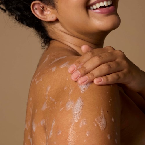En smilende kvinne med såpe og lotion på skulderen, som viser glatt hud og nyter en hudpleierutine.