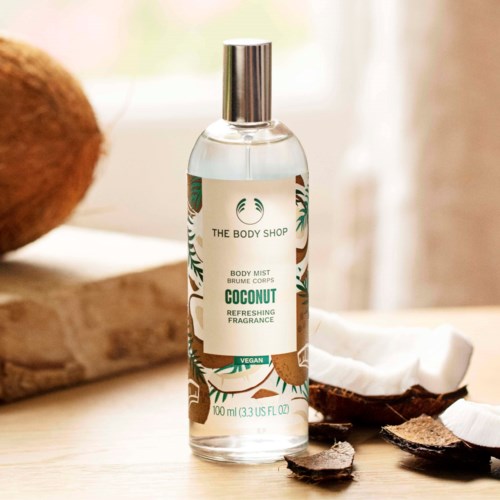 A transparent spray bottle labeled coconut body mist on a wooden surface, with coconut shells nearby and soft natural light.