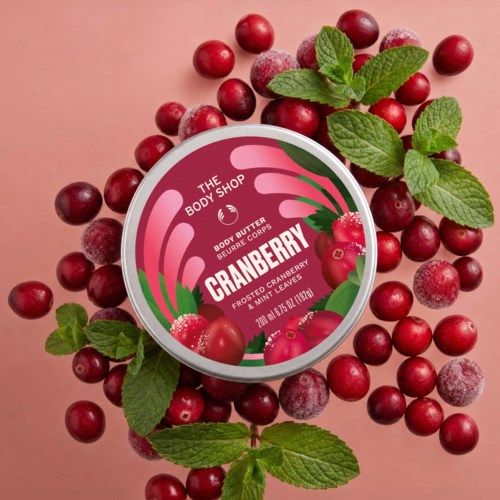 Kroppssmörbehållare med tranbärsetikett omgivet av färska och frostiga tranbär och myntablad på rosa bakgrund.