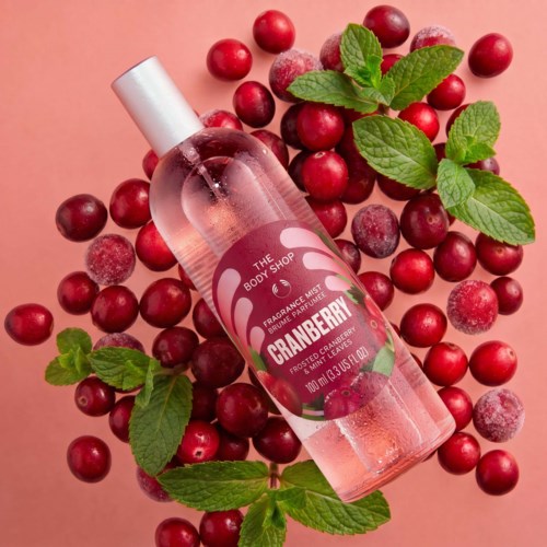Cranberry-scented body mist in a clear pink bottle surrounded by cranberries and green mint leaves.