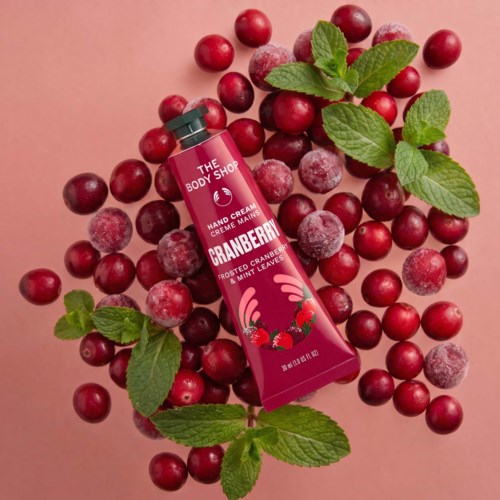 A tube of cranberry hand cream surrounded by fresh cranberries and mint leaves, on a pink background.