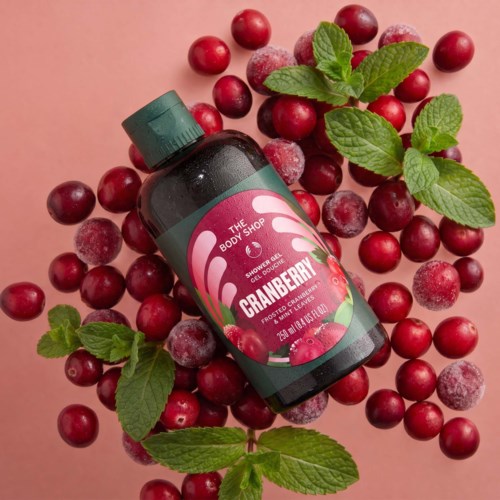 Cranberry duschtvålflaska omgiven av färska och frostiga tranbär med gröna myntablad på rosa bakgrund.