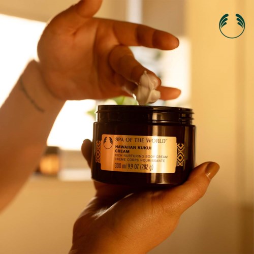 A person holds a black jar of Hawaiian Kukui cream, with some cream on fingers, in a cozy indoor setting.
