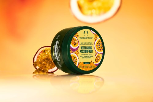 En grøn beholder fra The Body Shop passionfrugt scrub med passionsfrugt ved siden af, på en gul baggrund.