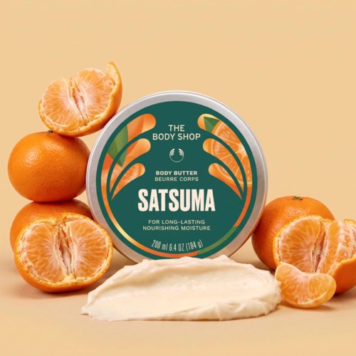 En boks body butter fra The Body Shop med hele og skrelte appelsiner rundt den og en klatt av smøret.