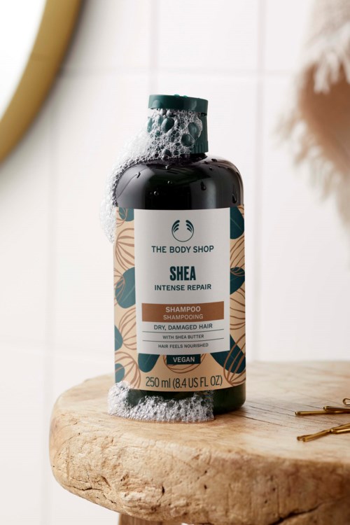 The Body Shopin Shea Intensive Repair -shampoo pullo, jonka päällä on vaahtoa, asetettuna puiselle pinnalle.
