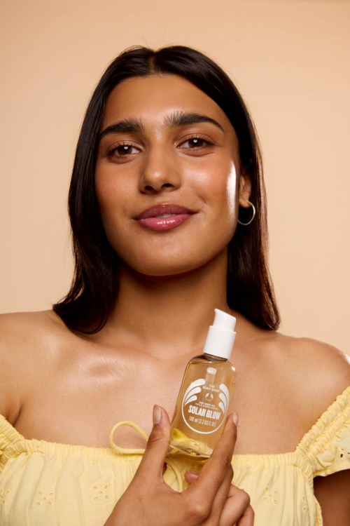 En kvinde med langt mørkt hår holder en lille flaske solcreme, smiler blidt mod en beige baggrund.