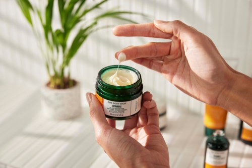Hænder holder en lille krukke med creme, med lidt creme der bliver skovlet ud. Der er en plante og andre flasker i baggrunden.