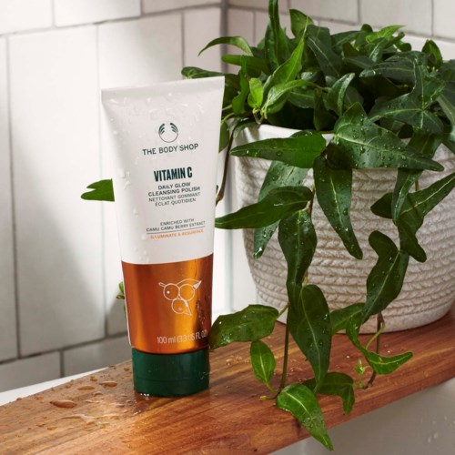 En hvit tube fra The Body Shop Vitamin M Bright Glow rens next to en grønn bladplante på en treflate.