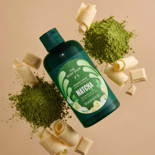 En grön flaska med duschkräm märkt The Body Shop Matcha, omgiven av matchapulver och vita chokladbitar.