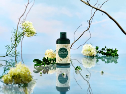 En flaske hvit te og elderflower lotion omgitt av blomster og grener, med blå himmel i bakgrunnen.